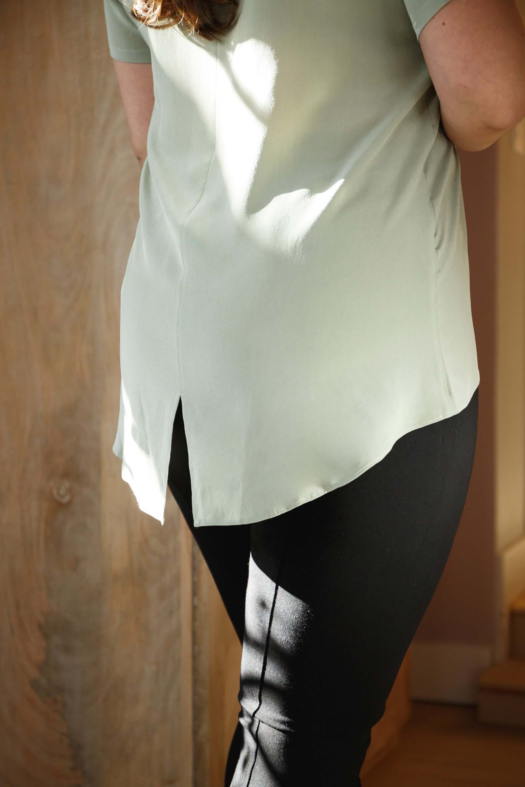 Woman wearing sage green silk tee top with relaxed fit and high low hem showing back view