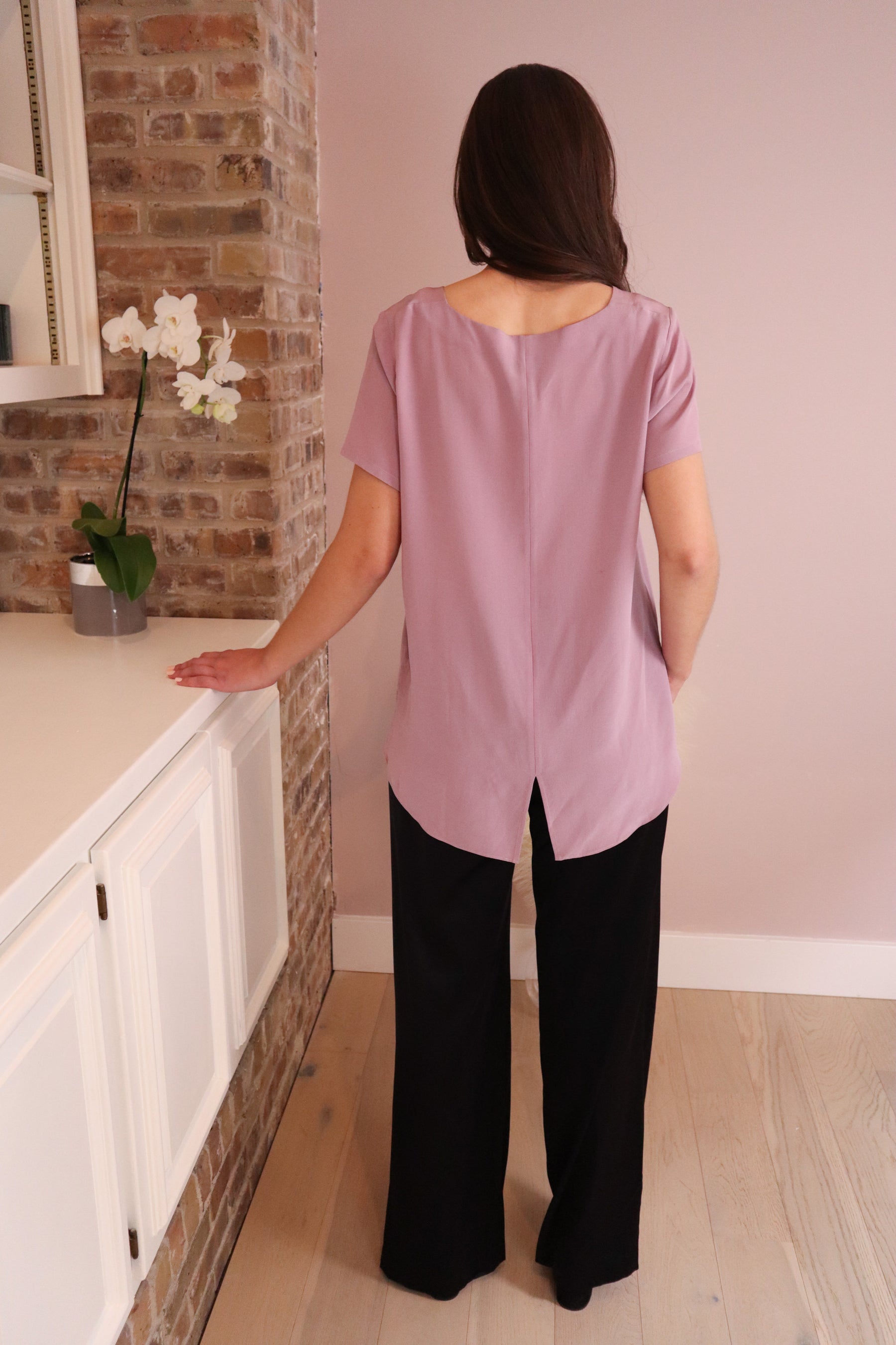 back view of a woman wearing lilac top untucked to show the high-low hem. She is also wearing a pair of black silk pants